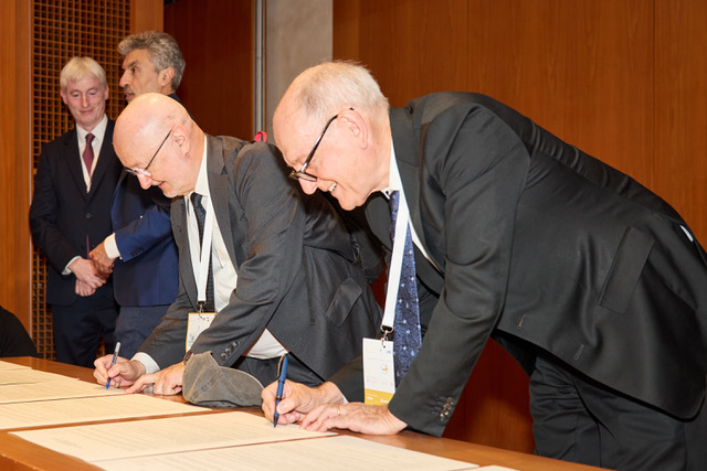Alex Waibel and another man, both in suits, bend down to sign documents laid out on a table in a room with wood paneled walls.