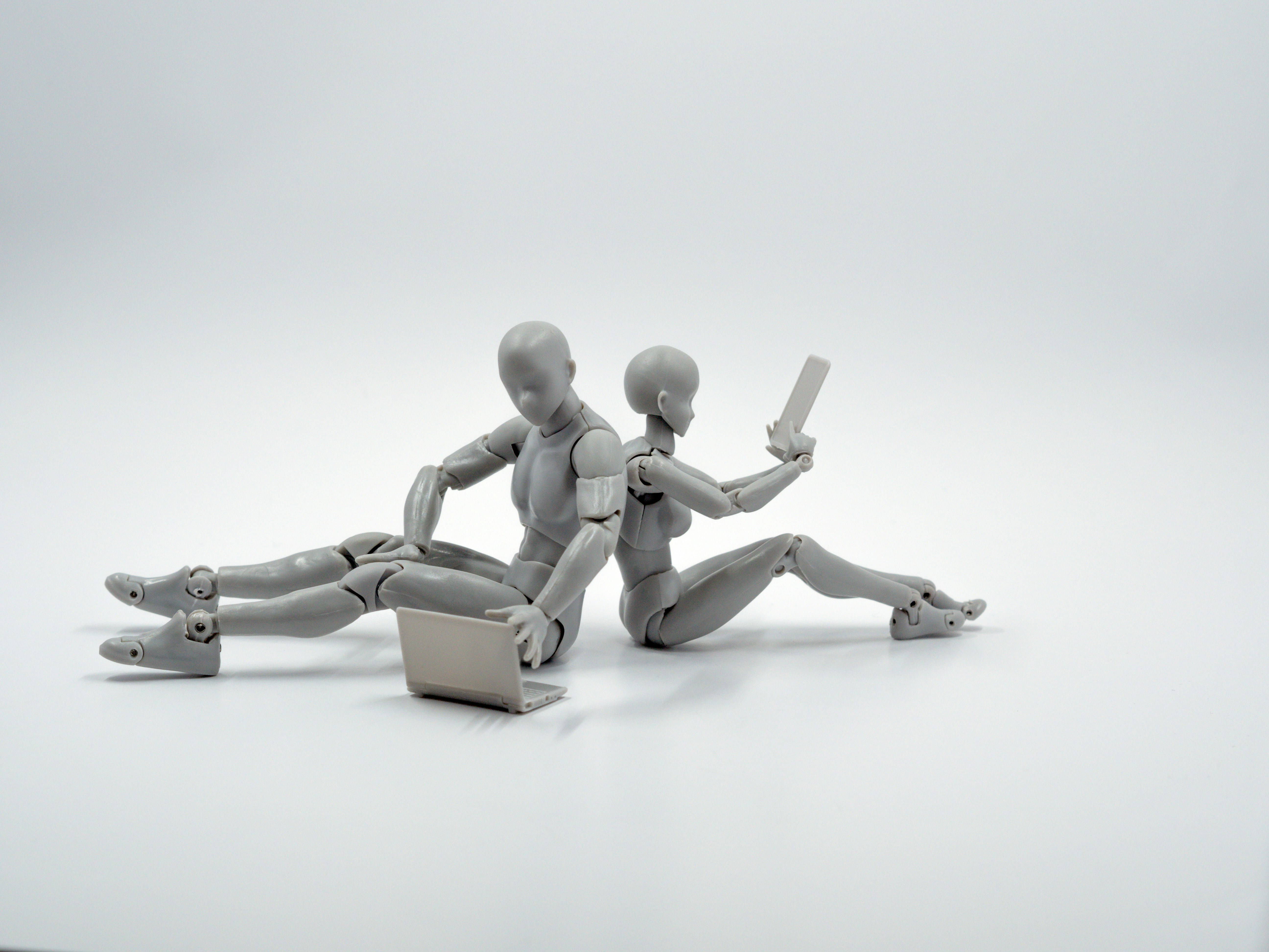 Two humanoid figures, possibly robots, sit on the ground, one holding a tablet and the other using a laptop