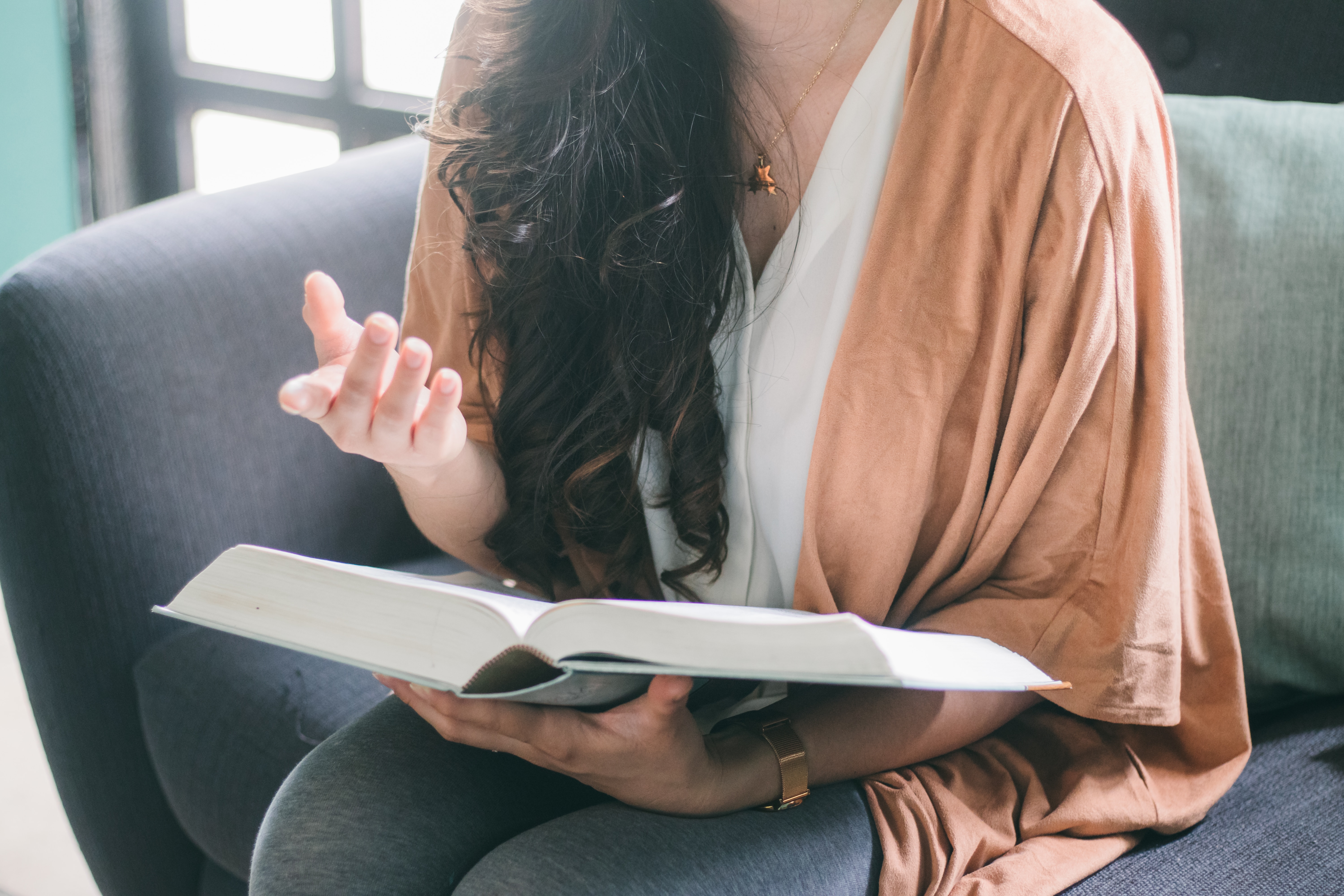 Stock photo of a person with long dark hair, visible only from the neck down, sitting in a chair and holding an open book with one hand and gesturing toward another an unseen person with the other.